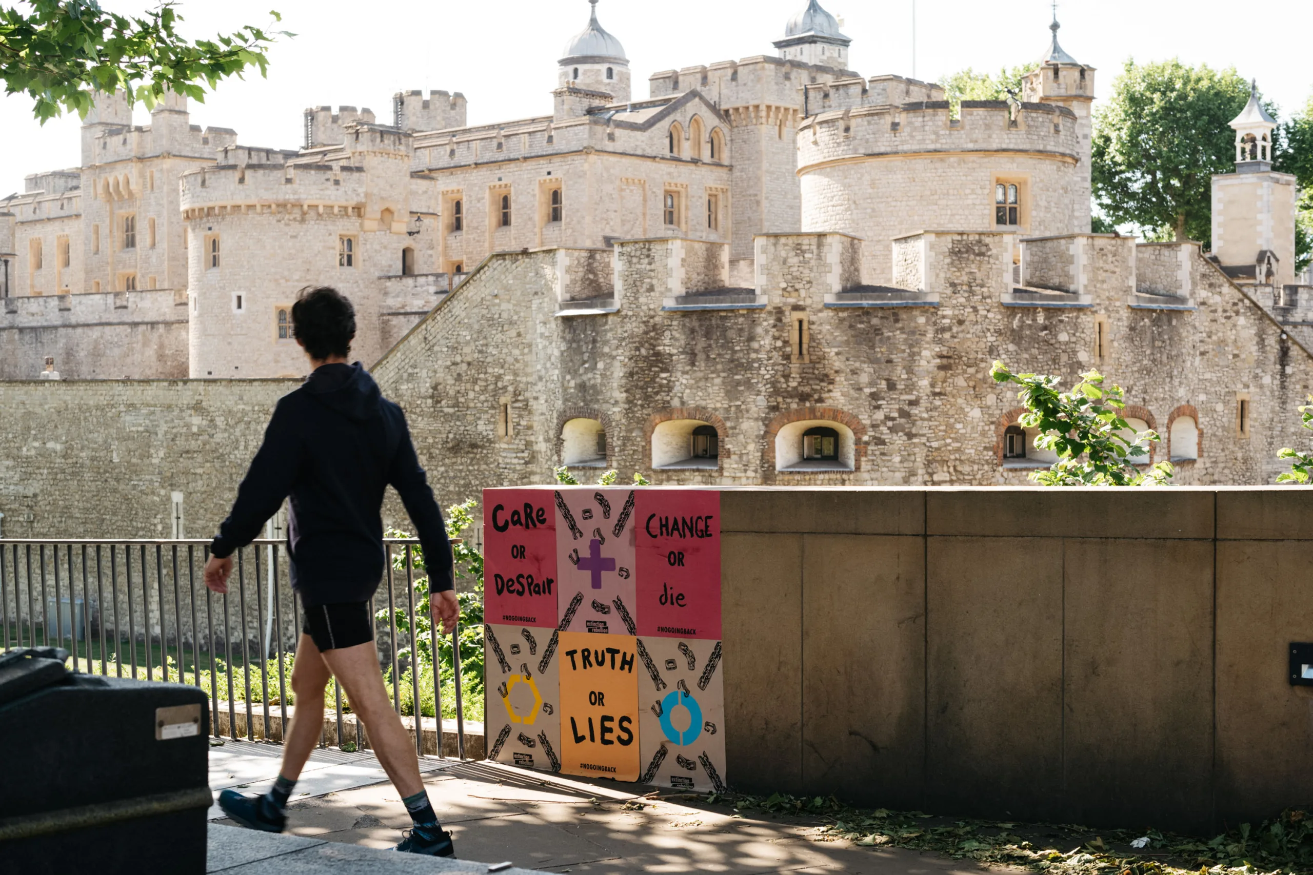 A person passing by a colorful protest sign with various messages, set against the backdrop of an ancient stone fortress with towers under a clear sky.