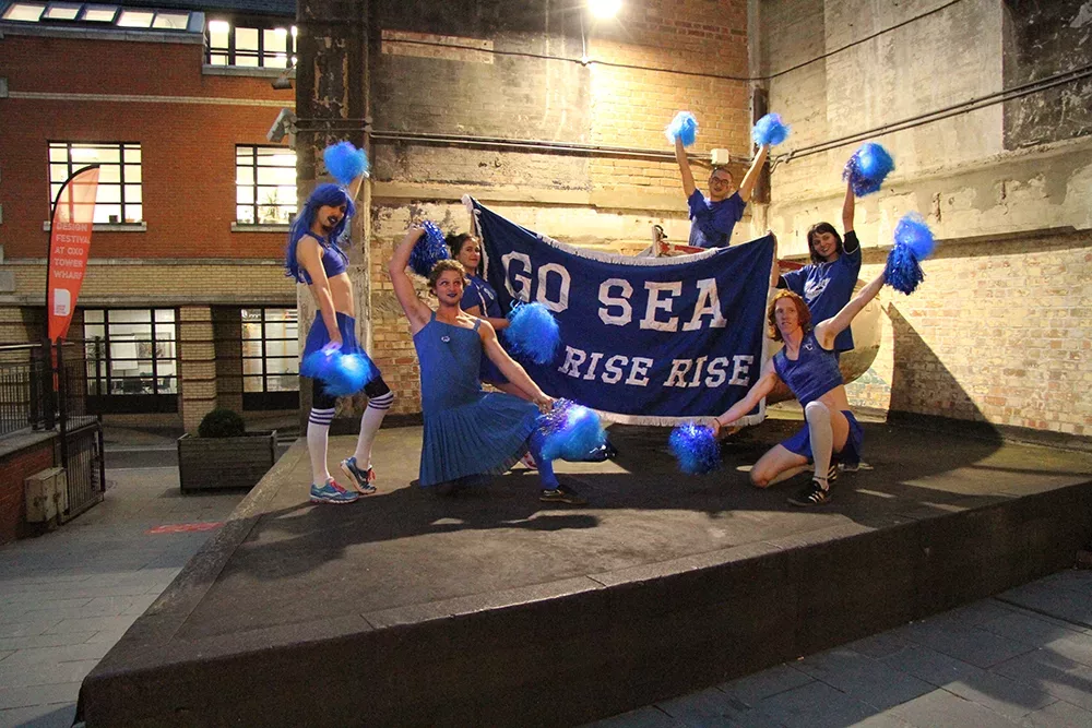 A group of people dressed in blue cheerleading outfits are holding pom poms and a banner that reads go sea rise rise rise
