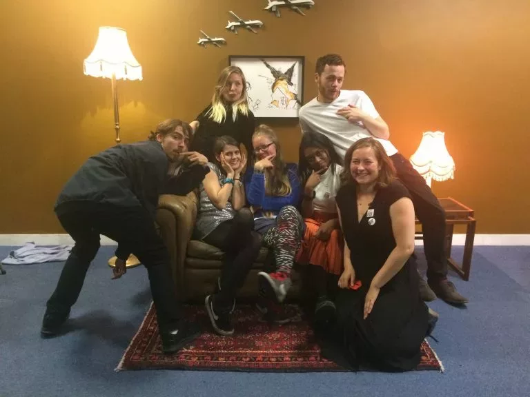 The art the arms fair team pose on Banksy's sofa installation with drones on the wall over a childrens drawing.