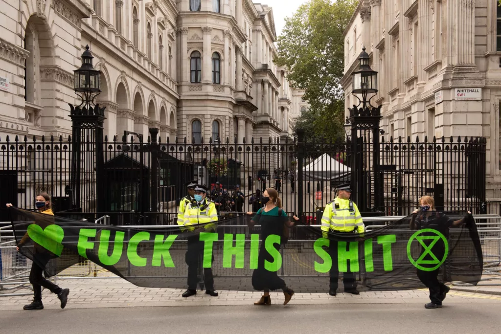 Protesters holding a banner with explicit language expressing frustration, in front of a gated building, as police officers observe.
