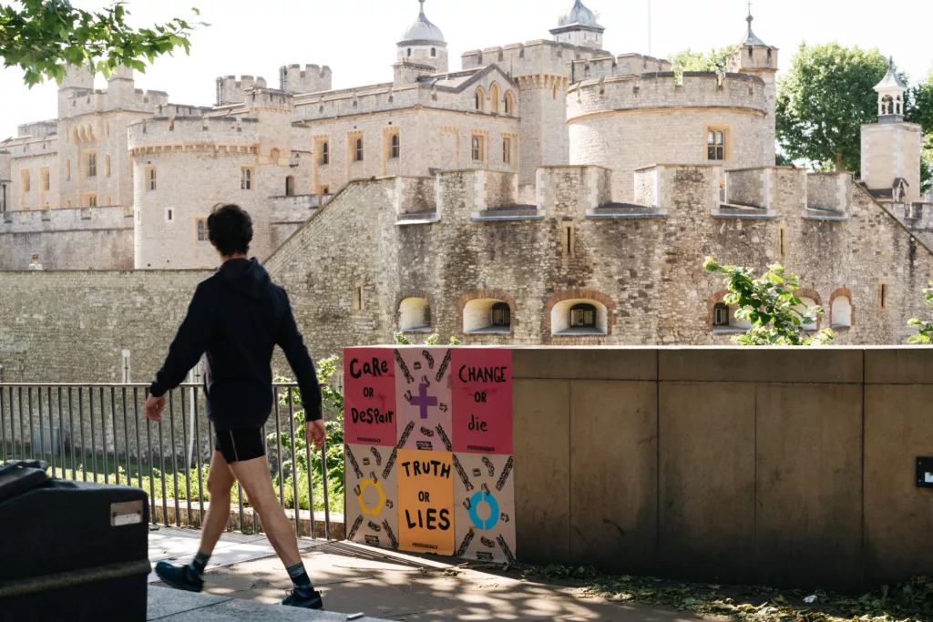 A person passing by a colorful protest sign with various messages, set against the backdrop of an ancient stone fortress with towers under a clear sky.