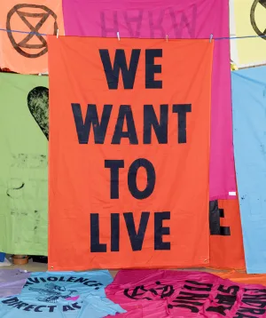 A vivid orange protest banner with bold black letters saying "we want to live" hangs amidst other colorful banners with various messages, symbolizing a call for action and the urgency of social or environmental issues.