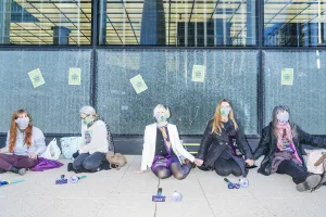 A group of activists engaged in a peaceful protest, wearing face masks and sashes, sitting in front of a building with windows splattered with green paint, symbolizing their commitment to environmental causes.